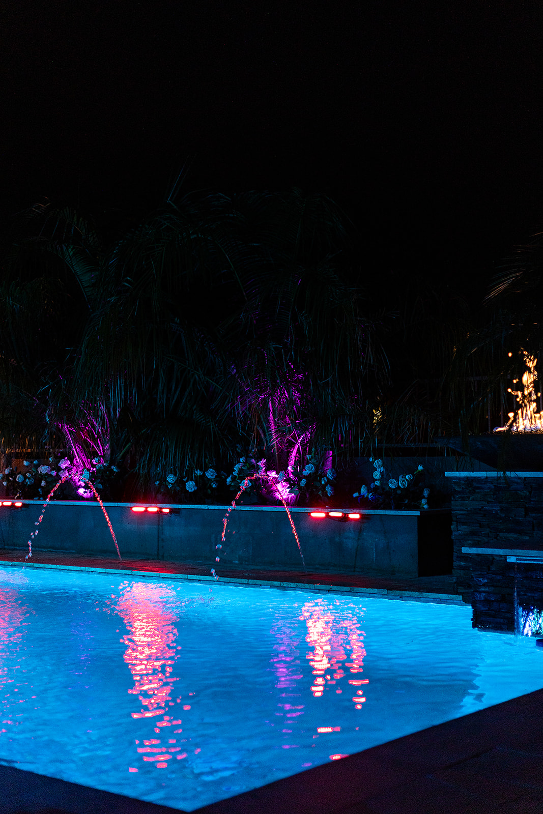 Blue illuminated swimming pool with arcing deck jets, pink accent lights, purple palm tree uplighting and a raised fire feature.