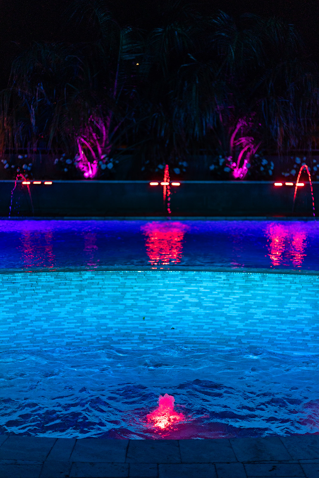Blue illuminated pool with a pink lit bubbler, arcing water jets and purple uplighting on palm trees.