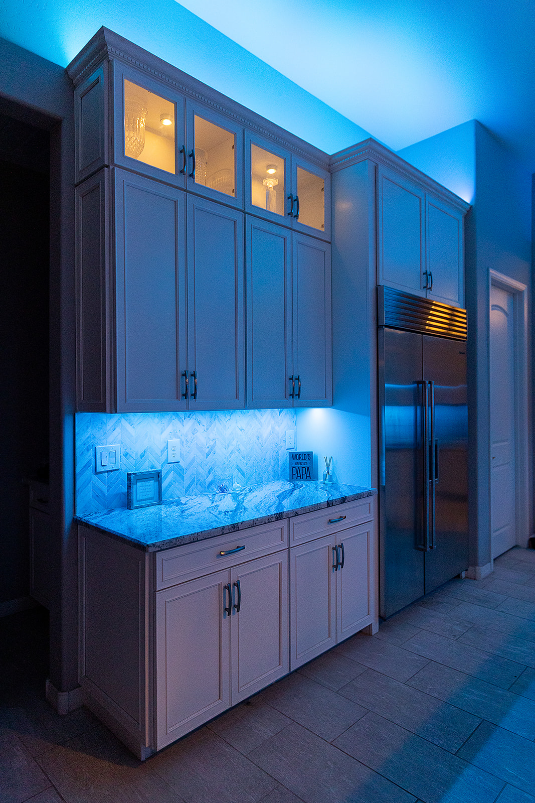White kitchen cabinetry with bright blue under cabinet lighting illuminating a herringbone backsplash and warm interior display cabinet lights.