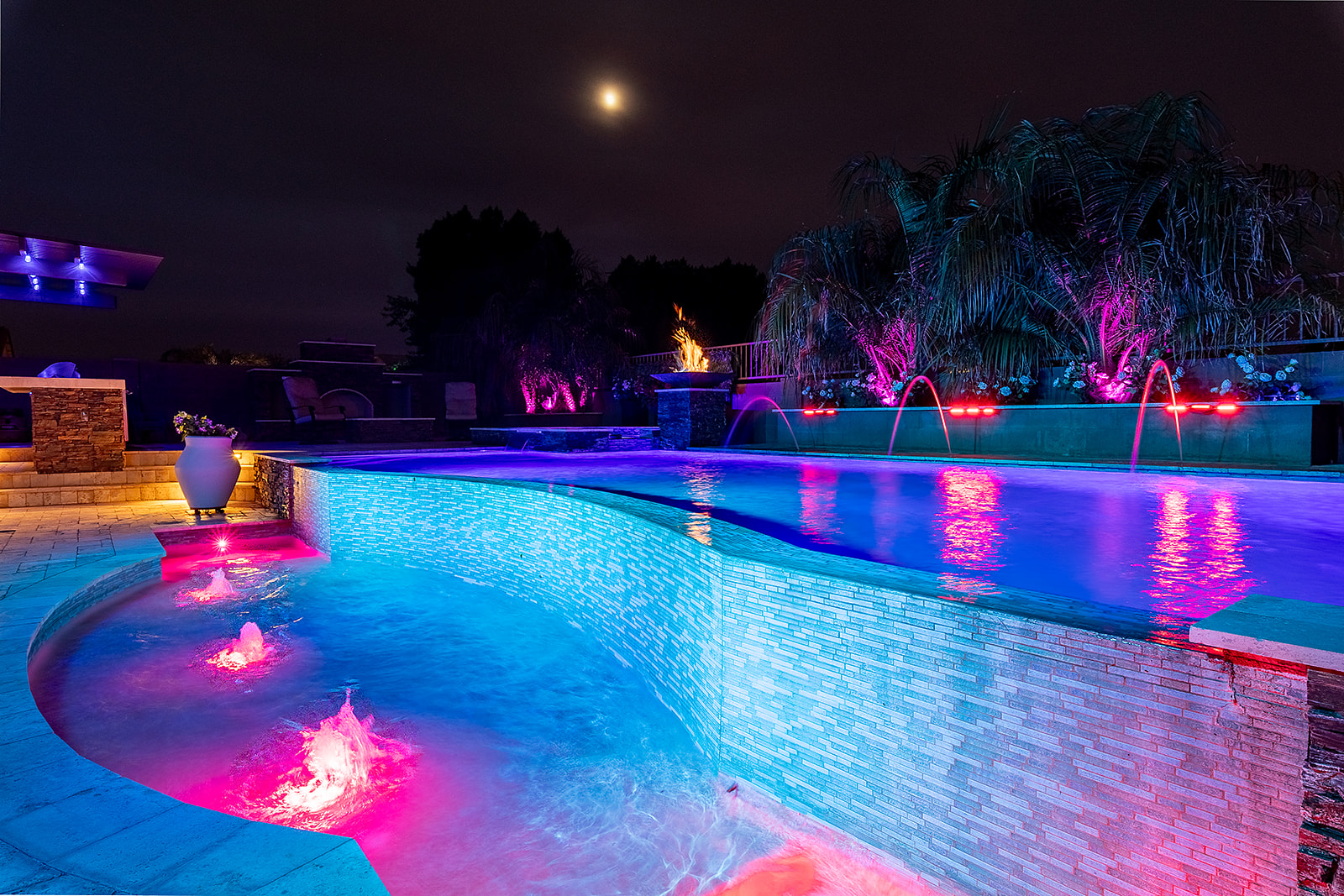 Luxury backyard pool illuminated with blue and pink LED lighting, lit water features, colorful palm tree uplighting and accent lighting.