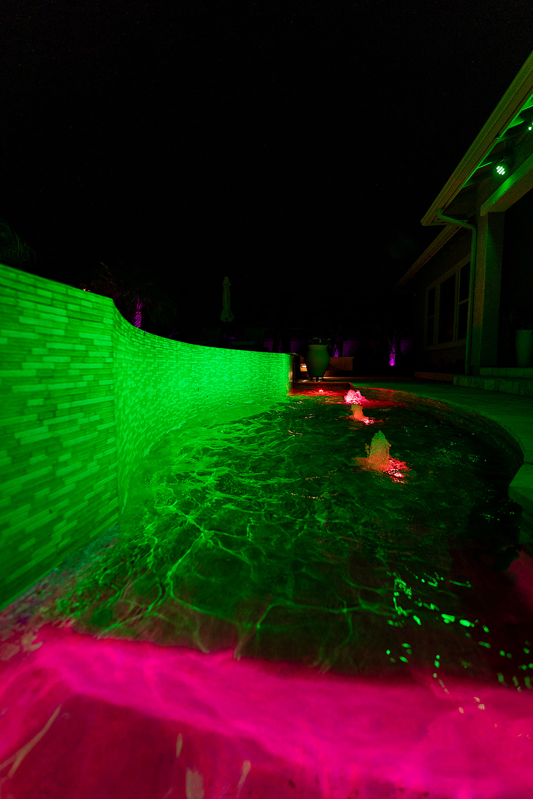 Close view of a pool illuminated in green with pink lit bubblers and colorful landscape accent lighting around the backyard.
