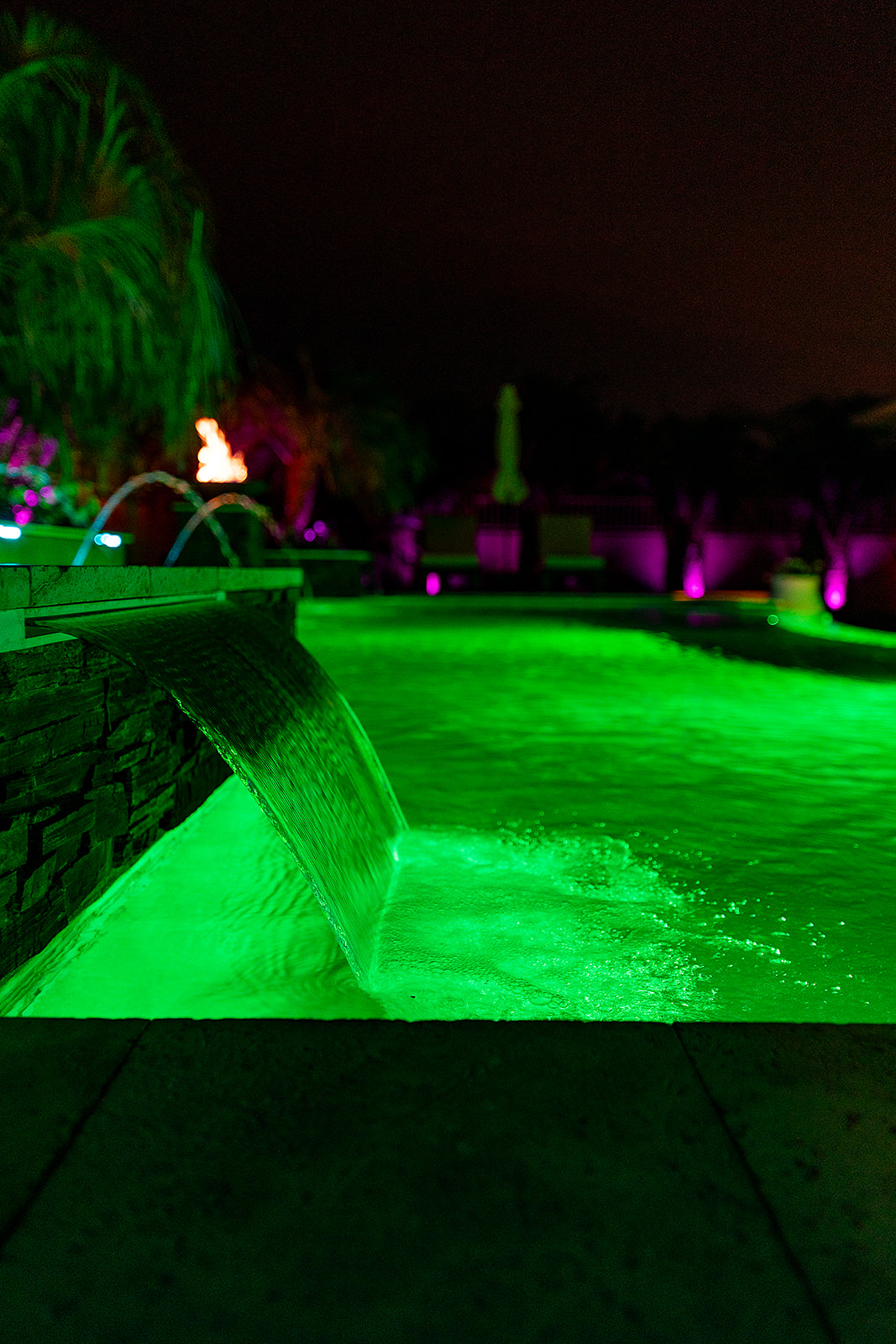 Close view of a sheet waterfall pouring into a swimming pool illuminated with vivid green LED lighting.