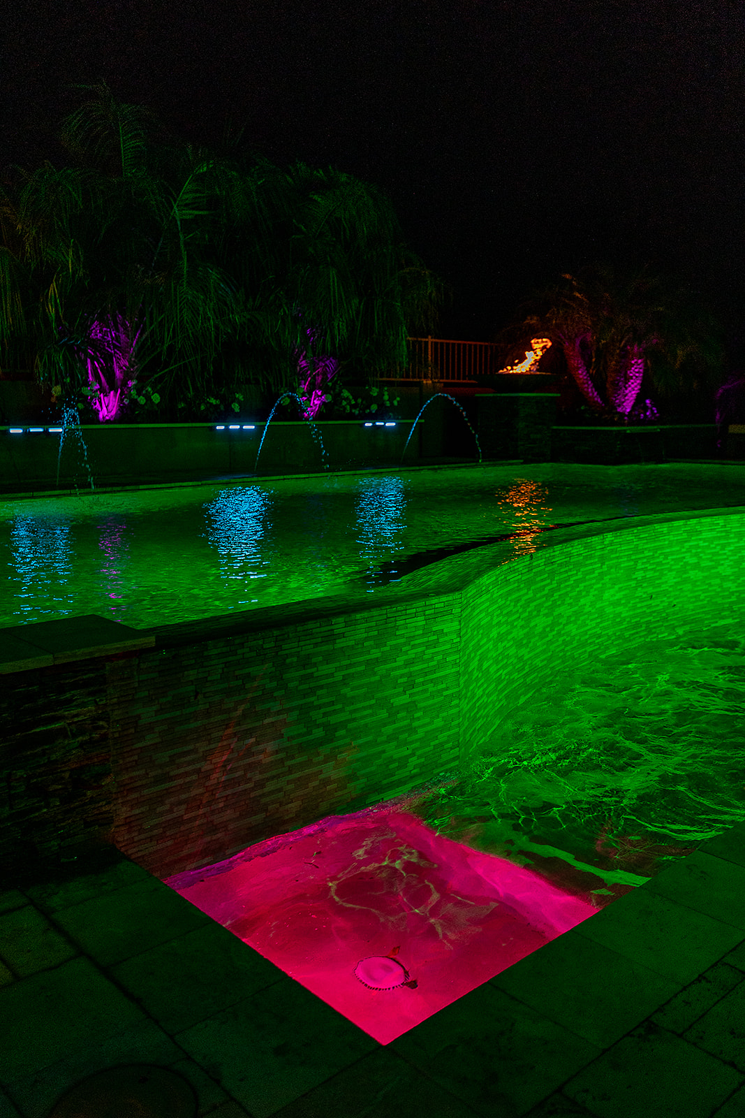 Nighttime pool with green underwater LED lighting, blue illuminated water streams, purple palm tree uplighting and a fire feature in the background.