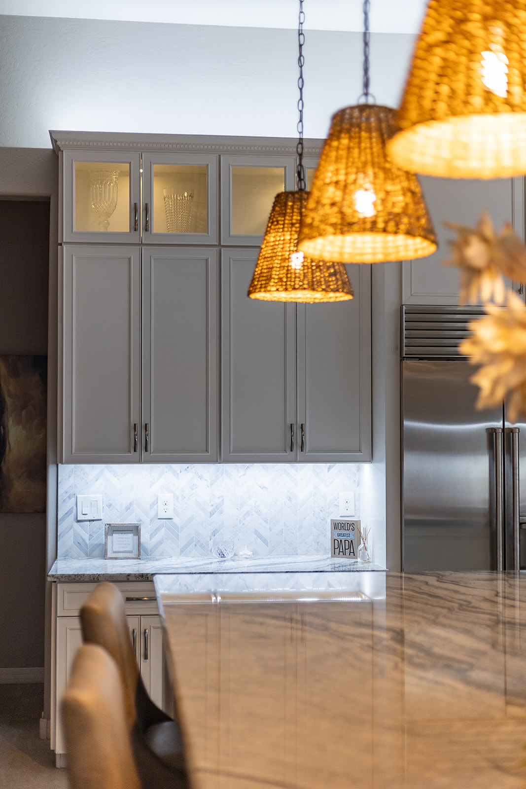 Warm woven pendant lights hang over a stone kitchen island with illuminated display cabinets and bright under cabinet backsplash lighting in the background.