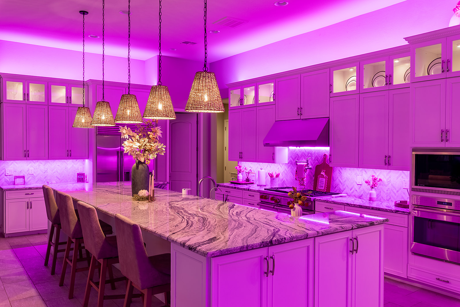 White custom kitchen illuminated by vivid pink LED cove lighting and under cabinet lighting with warm pendant fixtures over the island.