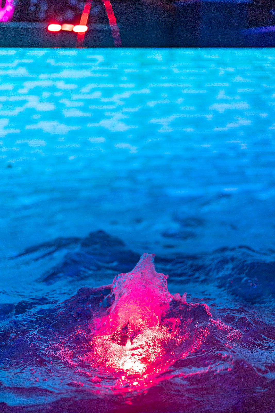Closeup of a pool bubbler glowing pink against blue illuminated water at night.