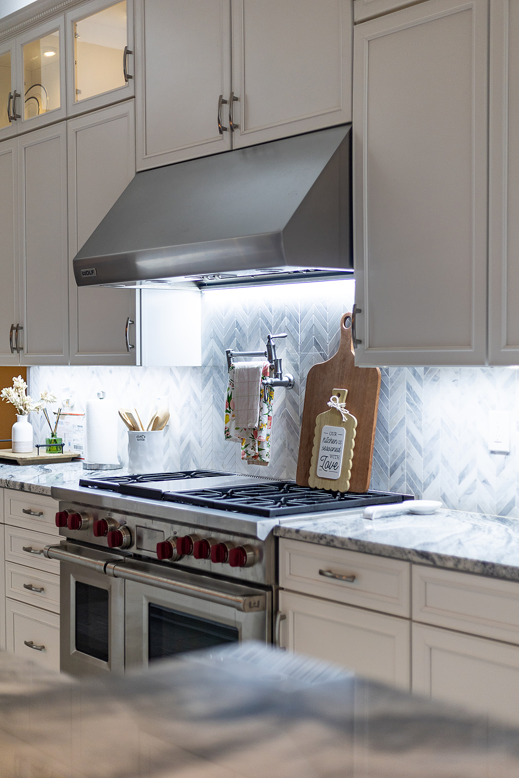 Professional style range beneath a stainless steel hood with bright task lighting illuminating the herringbone backsplash and surrounding counters.